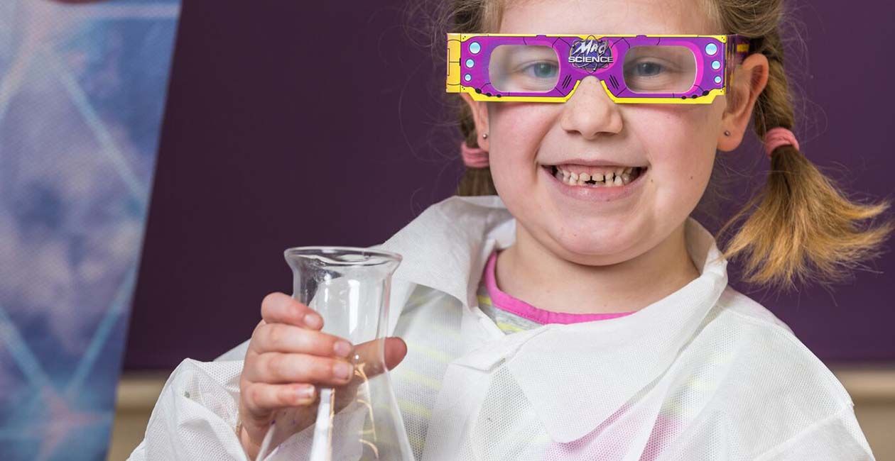Girl smiling in Mad science lab coat holding a beaker wearing purple diffraction glasses