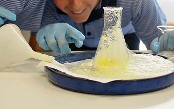 A boy's hand pointing at a beaker with yellow liquid overflowing onto a blue tray